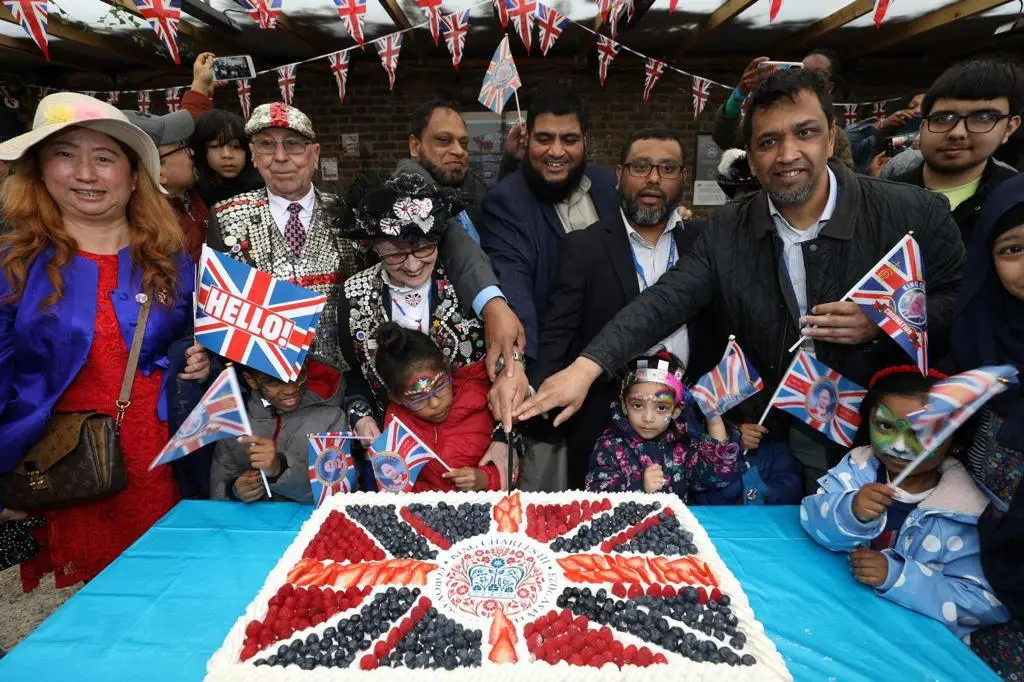 Large group of people cutting a cake decorated with strawberries, raspberries and blueberries in the style of a Union Jack flag. A Pearly King and Queen are among the group as well as some young children waving some special Coronation celebration flags.